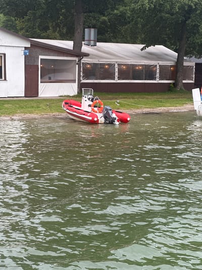 A red and white inflatable boat with a black outboard motor is tied to the shore of a body of water. The boat is positioned on the water's edge, with its bow facing left. A life preserver ring is visible on the boat's stern. In the background, there is a building with a white facade and a brown wooden door. Adjacent to this building is a covered outdoor seating area with clear plastic siding and lights inside. Trees and foliage form a dense green backdrop behind the structures. The scene is set on the shore of Lake Kłodno in Chmielno, Poland.