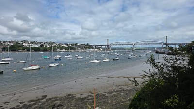 The image shows a wide, calm river or estuary filled with numerous sailboats and a few motorboats. The boats are moored in the water, indicating a harbor or marina. In the background, a large, modern suspension bridge spans the water, connecting two land masses. Along the hillsides of the far shore, a town or city with many houses and buildings is visible, nestled amongst trees. The sky is partly cloudy with a mix of gray and white clouds, suggesting a temperate day. The foreground shows a muddy shoreline with some vegetation and rocks. The overall scene conveys a sense of peaceful activity and a picturesque coastal or riverside setting. The location context of Plymouth, United Kingdom, aligns with the presence of a significant bridge over water and a coastal town. No people are clearly discernible in the image, and no text is visible.