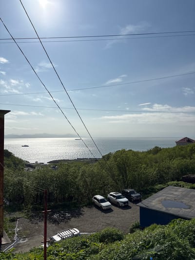 This media file captures an outdoor scene with a body of water, lush green trees, a few vehicles, and power lines against a bright sky. The sun is shining, creating a bright, clear day, likely daytime.
The scene appears to be a coastal or lakeside area with a wide expanse of water reflecting the sunlight. The water is calm, with a few small boats or ships visible in the distance. Dense green foliage, likely trees and bushes, borders the water and extends to the right side of the frame, suggesting a natural or semi-natural environment.
On the right side of the image, there's a paved area or driveway where three cars are parked. Two are white, one is a SUV, and one appears to be a sedan. Another white vehicle, possibly a van or truck with a rack on top, is parked further down. A dark, flat-roofed structure with a puddle on its surface is also visible, possibly a building or shed.
Dominating the left side of the frame are a series of power lines, crisscrossing the sky. The sky itself is a clear, bright blue with scattered white clouds. The overall mood is serene and peaceful, evoking a sense of a pleasant day outdoors.
There is no visible text in the image. The location context is Vulkanyy, Russia.