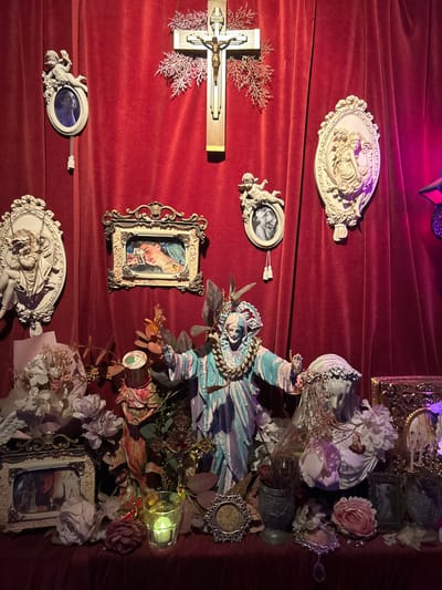 This image depicts an elaborately decorated display within a venue in 마포구, South Korea. A deep red velvet fabric serves as a backdrop, draping both a vertical wall surface and a horizontal surface resembling a shelf or altar.
Mounted centrally on the red velvet wall is a wooden crucifix featuring a metal corpus of Jesus, flanked by sprigs of artificial pink foliage. To its upper left, an oval, cream-colored ornate frame with cherubim holds a monochrome photographic portrait. Below this, a larger, cream-colored oval relief features classical figures. To the right of the crucifix, another oval, cream-colored ornate frame with cherubim holds a different monochrome photographic portrait. A large oval, cream-colored relief depicting classical figures is positioned to the right of this frame. A rectangular, ornate gold frame is situated below the crucifix, containing a painting of a woman with prominent eyebrows and a parrot, stylistically resembling Frida Kahlo.
On the red velvet-covered horizontal surface below, a collection of objects forms a complex arrangement. Prominently in the center is a large statue of a robed figure, painted in pastel blues, purples, and white, with arms outstretched and a golden wreath around its neck. To its immediate left, a smaller, multi-colored figurine is partially obscured by artificial foliage, which is interspersed throughout the entire display. Near the center, a clear glass votive holder contains a lit green candle.
To the left of the central statue, a cluster of artificial white and purple flowers is arranged next to an ornate silver-colored frame holding a photographic portrait. To the right of the central statue, a white bust of a veiled female figure, adorned with white artificial flowers and a sheer veil, stands prominently. Adjacent to it, an ornate dark metal urn contains dried foliage. Various smaller ornate frames, some containing images, and a small ornate silver-colored clock are scattered on the surface. A decorative pendant with a teardrop crystal also lies on the surface. The scene is illuminated with dim, atmospheric lighting, casting subtle shadows and highlighting the textures of the velvet and intricate decorations.