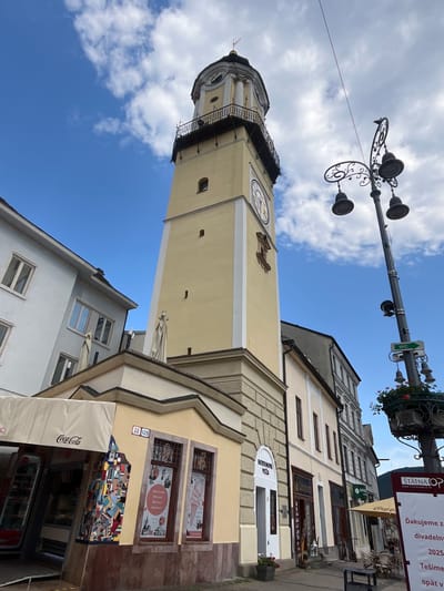 This media file is a vertical, eye-level shot of a street scene in Banská Bystrica, Slovakia, featuring a prominent clock tower and adjacent buildings. The sky is bright blue with scattered white clouds, suggesting a clear day. The clock tower, painted a pale yellow, has a dark balcony and a large clock face visible. Below the tower, storefronts are visible, with awnings and displays. One building has "HODINOVA VEŽA" written above an entrance, meaning "Clock Tower" in Slovak. Another sign indicates "ŠTÁTNA OPERA" (State Opera). Text on a poster advertises "ZMRZLINA, NAPOJE, WAFFLE, ZAKUSKY" (Ice Cream, Drinks, Waffles, Snacks). A Coca-Cola advertisement is also visible on an awning. A street lamp with decorative elements arches over the scene. The overall impression is of a sunny day in a historic European town square. The image appears to have been taken in the afternoon, given the sunlight.