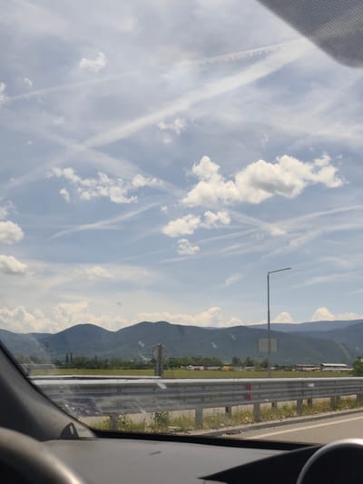 The image provides a view from the interior of a car, looking out through the windshield at a landscape near Skravena, Bulgaria, on a bright, sunny day. Parts of the car's dark dashboard and steering wheel are visible in the lower foreground.

Across the mid-ground, a multi-bar metal guardrail runs horizontally, separating the viewer's immediate space from the scene beyond. Faint, obscured text or stickers are visible on the guardrail, with one section appearing to contain a number sequence like "610701" or "610791."

Beyond the guardrail lies a flat, green expanse of land, which could be fields or an industrial area, featuring several low-rise buildings and structures in the distance, including what might be a chimney or tower on the left side. A tall, gray street lamp post stands prominently on the right side of the frame, reaching high into the sky.

In the background, a range of dark, rolling mountains extends across the horizon. The sky dominates the upper portion of the image, displaying a clear, light blue color dotted with scattered white cumulus clouds. A notable feature of the sky is the presence of numerous airplane contrails, forming distinct white lines and crisscrossing patterns. The image depicts a calm, daylight scene, suggesting travel or a scenic stop.