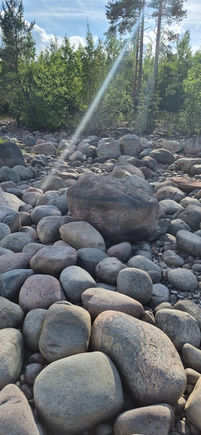 The image depicts a rocky shore with various sized stones and boulders. In the background, there is a dense forest with green trees. The sky is blue with some white clouds, and a sunbeam is visible, creating a lens flare effect. The scene appears to be outdoors in a natural setting, likely near a body of water, given the presence of smooth stones. The location is specified as Motornoe, Russia. There are no people or animals visible in the image, and no actions or interactions are taking place. The focus is on the natural landscape and the geological features of the stones.