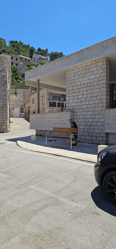 An outdoor, daytime scene captures a person on a bench in front of a stone building. The person, appearing elderly with gray hair, is wearing a black t-shirt and is leaning forward, looking down at something on the bench. The bench itself is made of wood with metal supports. To the right of the bench, a portion of a black car is visible, parked on the street. The environment includes a paved road with white lines, a sidewalk, and a building constructed from light-colored bricks. In the background, more stone buildings and lush green trees are visible on a hillside under a clear blue sky. The location is in Općina Pučišća, Croatia.