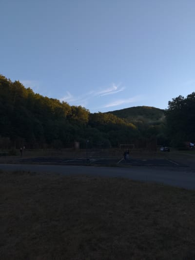 A medium shot captures an outdoor scene with a clear blue sky above a wooded hill. The hill is densely covered in dark green trees, with some areas illuminated by sunlight, creating a contrast between shadow and light. A few wispy clouds are visible in the sky. In the foreground, a dark asphalt surface marks what appears to be a sports court or parking area, with faint lines indicating boundaries. A grassy area with sparse vegetation is in the immediate foreground, mostly in shadow. Two people are standing on the dark surface, one appearing to be walking. Further back, there are some structures, including a net that could be part of a volleyball court, and a fence. In the distance, a few vehicles are parked. The overall impression is of a somewhat secluded outdoor recreational area, possibly part of a park or community space, situated at the base of a hilly, forested landscape.