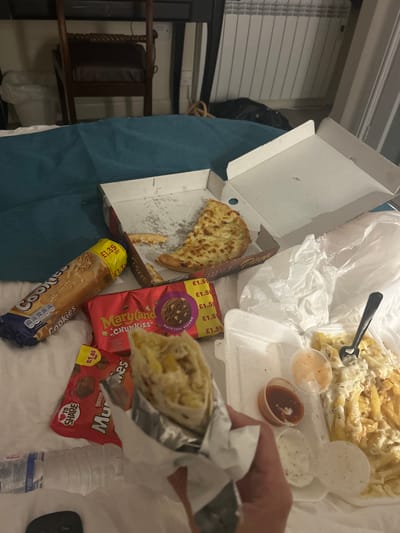 The image shows a collection of takeaway food and snacks arranged on a bed.

**Food:**
*   A partially eaten pizza in a pizza box.
*   Chips covered in sauce on a white polystyrene tray.
*   Small pots of sauces.
*   A wrap or burrito held in a hand.

**Snacks:**
*   Maryland Chunkies cookies.
*   Nestle Munchies.
*   A McVitie's Cookie bar.

**Other:**
*   A plastic fork.
*   A bottle of water.
*   A television remote

**Environment:**
*   The setting appears to be a bedroom, indicated by the bed.
*   The bedsheets are teal.
*   A dark chair can be seen near the bed.
*   A window with blinds is visible in the background.

There is no information available to confirm the image was taken in Aylesbury, United Kingdom.