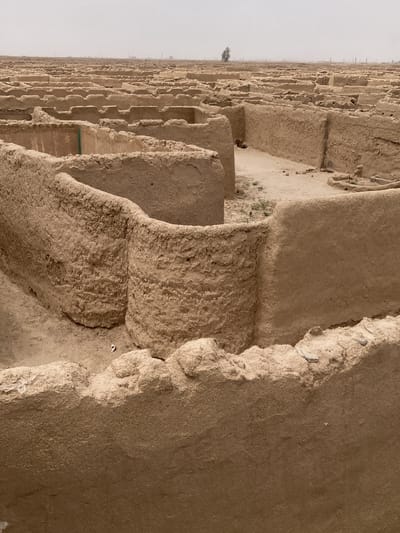 The image shows the ruins of Derawar Fort, located near the city of Saranan in Pakistan. The scene depicts an extensive network of interconnected structures with crumbling mud walls, forming a maze-like pattern. The architecture is characterized by rounded and rectangular shapes. The ground between the structures is sandy, with sparse patches of vegetation in some areas. In the background, the landscape is flat and hazy, with a single tree visible in the distance. The overall impression is one of an ancient and desolate settlement.