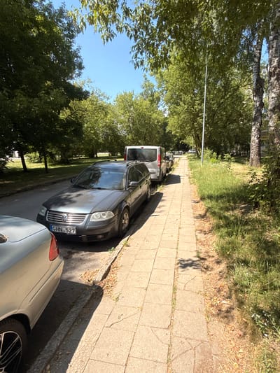 A car with a license plate reading "GHN 527" is parked on the side of a street in Rudamina, Lithuania. The car is dark gray and appears to be a station wagon. It is parked next to a sidewalk made of concrete pavers. To the left of the car, a portion of a silver vehicle is visible. Behind the gray car, there are other vehicles and a white van. The street is lined with trees, and there is a grassy area with some sparse vegetation and scattered leaves on the right side of the sidewalk. The sky is clear and blue.