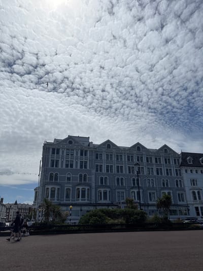 The image depicts a building in Llandudno Junction, United Kingdom, with a light gray facade and multiple windows. The building has a distinctive stepped roofline. Lush greenery and palm trees are situated near the building's base. A paved walkway runs parallel to the building, and blurred figures of people can be discerned along the walkway. Overhead, a sky filled with cumulus clouds covers most of the frame. A single bird flies in the direction of the building.
