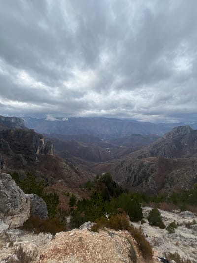 This image depicts a wide panoramic view of a rugged mountainous landscape in the city of Klobuk, Bosnia and Herzegovina, under an overcast sky. The foreground consists of light-colored rocky terrain and sparse, low-lying vegetation, including small evergreen trees and shrubs, sloping downwards from the vantage point. The midground features a vast expanse of valleys and multiple mountain ranges, primarily covered with dormant, brownish vegetation and prominent grey rock formations. A distinct, large vertical rock face is visible on the left side of the midground. In the deeper valleys, a winding path or road can be observed. The background reveals further distant mountain peaks, some partially snow-capped, partially obscured by atmospheric haze or low clouds. The sky is entirely covered with dense, grey clouds, indicating an overcast weather condition. No people, animals, or immediate human structures are visible.