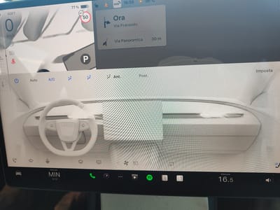 The image displays the interior of a Tesla vehicle, specifically focusing on the central touchscreen and dashboard. The touchscreen shows navigation information, with "Ora" and "Via Prarondo" displayed, indicating a destination in Orta San Giulio, Italy. The speed is displayed as "0 km/h" and the battery charge is "77%". The climate control interface is visible, showing fan speed set to "10" and a temperature of "16.5". The steering wheel is also visible. The overall environment is that of a parked vehicle's interior.