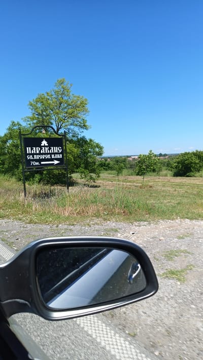 The image shows a black road sign in Seltsi, Bulgaria, indicating "Paraklis Sv. Prorok Iliya 70m" (Chapel of Saint Prophet Elijah, 70 meters). The sign features a small depiction of a church at the top and an arrow pointing right. Behind the sign are trees, tall grass, and a distant view of a settlement with red-roofed buildings. The clear, blue sky dominates the upper half of the frame. The lower part of the image features the side mirror of a vehicle, suggesting the photograph was taken from inside a car. The environment suggests a rural or semi-rural setting on a sunny day. No people are visible.