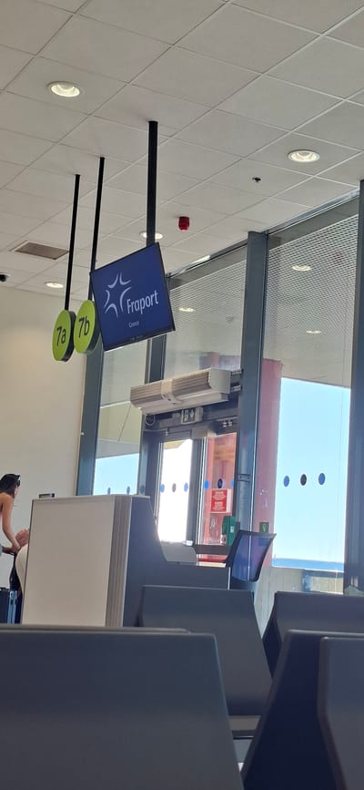 The image depicts an indoor scene, likely an airport or public waiting area, in Rhodes, Greece. The orientation of the photo is rotated 90 degrees counter-clockwise.
The primary subject is a sign with the "Fraport Greece" logo and the word "Greece" displayed on a dark blue screen. Below this screen, two circular green signs, labeled "7a" and "7b," are visible. A black pole supports the sign.
In the background, a large window or glass partition offers a view of the outdoors, suggesting a bright, sunny day. The sky is visible through the glass, with some reflections of the interior. The scene includes elements typical of an airport, such as seating areas (partially visible as grey structures on the right) and lighting fixtures on a white, diamond-patterned ceiling. A red fire alarm is mounted on the ceiling to the left of the sign.
The lighting suggests it is daytime, and the bright outdoor light indicates clear weather. A small section of a person's arm and shoulder is visible in the lower right corner, reaching towards something out of frame. No specific activity or event is clearly depicted, other than the general context of a waiting area.
Visible text includes "Fraport," "Greece," "7a," and "7b." There is also some text on a red warning sign visible through the glass, but it is too small to be legible.