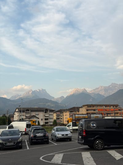 This is a vertically oriented photograph taken in Monthey, Switzerland, depicting a street scene with vehicles, buildings, and mountains in the background. The foreground shows a paved road with white markings, indicating a parking lot or street. Several cars are parked, including a dark-colored mini cooper with license plate "VS-609300" and a grey Volkswagen with license plate "VS-596716". A black van, possibly a commercial vehicle, is also prominently featured with a circular logo on its side. Behind the vehicles, there are several multi-story buildings with balconies and architectural details typical of European towns. The background is dominated by a majestic mountain range, partially covered in snow in some areas, under a sky with scattered clouds. The lighting suggests it is daytime, likely late afternoon, with soft, diffused light. The overall atmosphere is tranquil and scenic, highlighting the coexistence of urban infrastructure and natural beauty. Visible text includes license plate numbers and company names like "BENTSCHI AG DURRENASCH".