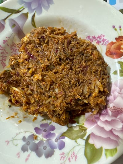 A mound of shredded, savory food rests on a floral-patterned plate. The food appears to be a mix of dark brown and tan, with visible strands of shredded ingredients. Red onion pieces and light-colored slivers add texture. The dish has a moist or oily sheen. The plate is adorned with various flowers in shades of pink, purple, and orange.