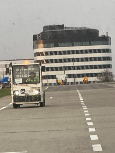 The image presents an outdoor scene at Budapest Ferenc Liszt International Airport, situated near Vecsés, Hungary. An overcast, light-grey sky dominates the background. In the foreground, a white ground support utility vehicle, branded with "MENZIES AVIATION" and "HISERV", moves forward on an asphalt service road delineated by a dashed white centerline. The vehicle's two front headlights are illuminated, and a single amber beacon flashes on its roof. A solitary individual occupies the driver's seat, clad in a dark head covering, ear protection, and a lime green and black high-visibility jacket. In the mid-ground, to the vehicle's left, a portion of a blue tarp or tent is visible. The background features a large, multi-story building characterized by a dark, curved upper section and lower floors with alternating horizontal bands of white and dark material. A partial "ONE" sign is affixed to the building's upper-left facade. Stationary on the tarmac between the moving vehicle and the distant building are two pieces of yellow airport ground equipment and a white utility trailer. A small, sparse tree or shrub is positioned to the right of this equipment.