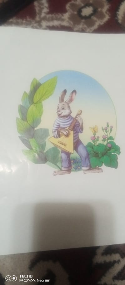 The image is a drawing of a cartoon rabbit playing a balalaika. The rabbit is wearing a striped shirt and blue pants. It is standing in a grassy area with some flowers and a butterfly. The background is a light blue sky. The drawing appears to be on a white piece of paper, which is placed on a surface with a patterned fabric. The bottom left corner of the image shows a "TECNO POVA Neo 2" logo, indicating the device used to capture the photo. The location of Tahmach, Uzbekistan, is not discernible from the image itself.
