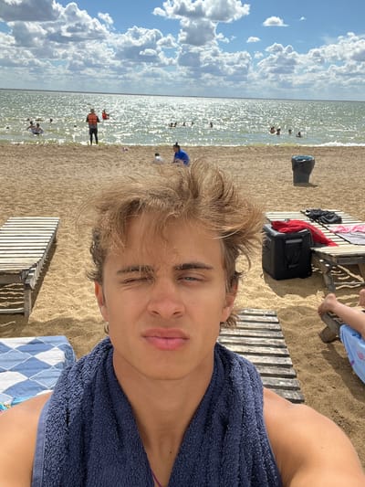 The image is a selfie taken at a beach, likely in the city of Gres-2, Kazakhstan. The foreground features a young man with messy blonde hair, winking at the camera, with a blue towel draped around his neck. Behind him, a sandy beach stretches towards the shimmering water of a lake or sea. Several people are visible in the water, swimming and enjoying themselves. Beach chairs and a trash can are also present on the sand. The sky is partly cloudy with bright sunlight reflecting off the water.