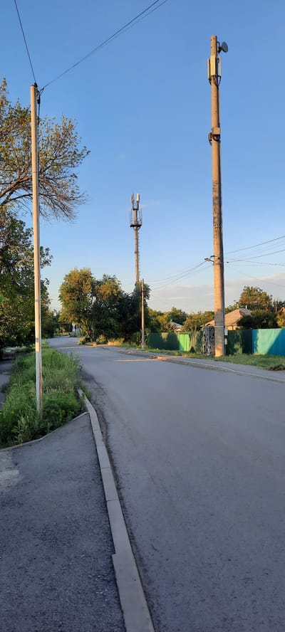 A paved road stretches into the distance under a clear blue sky in a semi-urban or rural setting. On the right side of the road, a tall, concrete utility pole stands, supporting electrical wires and a cellular tower. To its left, a slightly shorter cellular tower is visible, also equipped with antennas. Further left, another utility pole with wires is present. The roadside features a strip of green grass and vegetation. On the right side of the road, beyond a green fence, houses are visible. The sun is casting long shadows, suggesting it is either early morning or late afternoon. The overall scene depicts a street with infrastructure for utilities and communication. This scene is likely in Shakhty City District, Russia, based on its characteristic utility poles and housing styles. There are no people or vehicles visible in the image.
