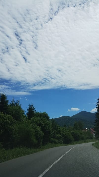 An asphalt road curves to the right through a green, mountainous landscape under a bright blue sky dotted with wispy white clouds. Dense green trees line the left side of the road, with a glimpse of a red-roofed house peeking through them in the distance to the right. A steep, tree-covered mountain dominates the background. The road has a white line down the center. The image appears to be taken from a moving vehicle.