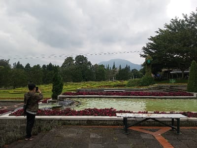 A person in a camouflage jacket takes a picture with a smartphone. A multi-tiered fountain with red flowers lines the edges. A bench sits in the foreground on a stone patio. Green lawns and trees are in the distance, with a mountain faintly visible. The sky is overcast, suggesting a cloudy day.
