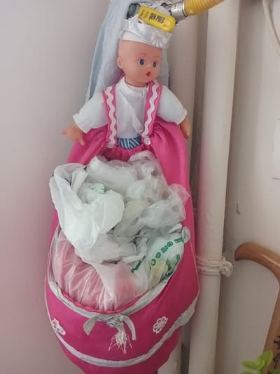 A doll, dressed in a pink and white outfit and a silver cap, hangs on a wall in Bahşılı Belediyesi, Turkey. The doll's dress serves as a storage pouch for several plastic bags. The doll is positioned to the left of a vertical pipe, which is wrapped with a white rope. A light blue cloth is draped behind the doll. A wooden cane handle is visible in the lower right corner of the frame. The wall appears to be a pale pink color.