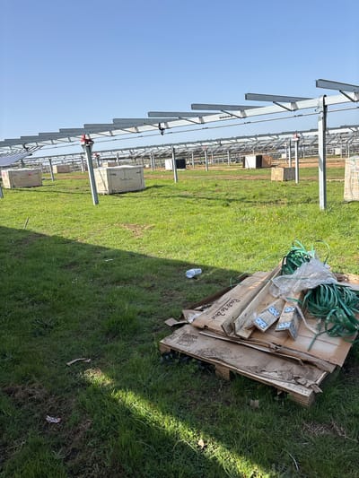 The image shows a landscape in Galatro, Italy, where solar panel installation is underway. In the foreground, there is construction debris including a wooden pallet with green strapping on top. The pallet has the text "Route to Zero" and "Committed to 100% Renewable" printed on it. Beyond the debris, the landscape is grassy, and the skeletal frames for solar panels stretch into the distance. Some of the solar panels have already been installed on the frames. The sky is clear and blue.