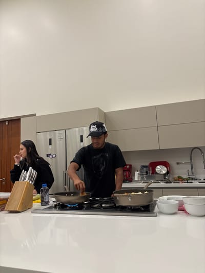 A man wearing a black t-shirt and a black cap is cooking in a modern kitchen. He is holding a large pan over a lit gas stovetop. Next to him, a woman in black is standing at the counter. A knife block and a water bottle sit on the counter nearby. Behind them is a refrigerator. The location is Santiago Etla, Mexico.