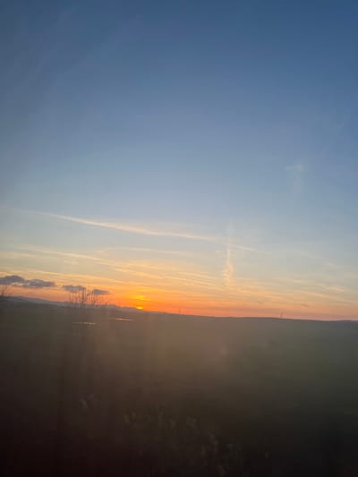 The image depicts a sunset over a rural landscape in or near Staro Oriahovo, Bulgaria. The upper two-thirds of the frame consist of a clear blue sky, transitioning to a vibrant orange and yellow gradient at the horizon where the sun is partially visible as a bright orb, positioned slightly left of center. Multiple linear contrails are present across the sky, with one prominent vertical contrail on the right. The horizon is defined by a dark, undulating line. In the midground, silhouetted against the sunset on the left, are several bare deciduous trees and a small, dark, reflective body of water. Distant hills or low mountains are visible on the far left. Two slender, vertical structures, likely power transmission towers, are faintly discernible on the right side of the horizon. The foreground is dark and indistinct due to the setting sun's glare, appearing as an expanse of shadowed land. No individuals or specific actions are discernible. The overall environment is open and natural, characteristic of agricultural or undeveloped land.