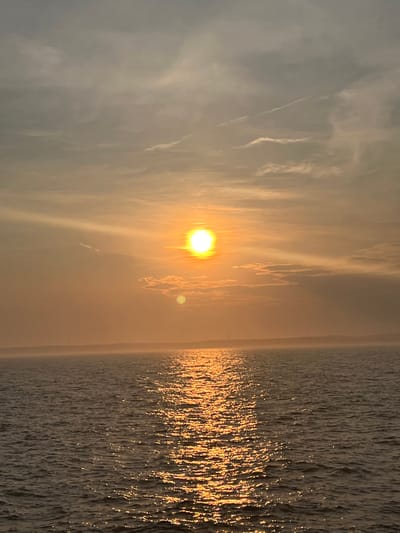 The image depicts a sunset over a body of water, likely the Saint John River in Saint John, Canada. The sun, a bright orb, is positioned in the upper center of the frame, casting a warm, golden light across the sky and water. The sky is a gradient of orange, yellow, and grey, with wispy clouds scattered throughout. The water is dark and rippling, with a shimmering reflection of the sun creating a path of light across its surface. In the distance, along the horizon, a faint landmass is visible, possibly with some structures or wind turbines. There are no people or specific objects that would indicate a precise location within the city, but the scene is characteristic of a waterfront view during sunset. The overall impression is one of natural beauty and tranquility.