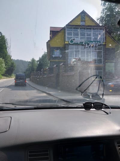 This is a daytime, eye-level, exterior shot from inside a vehicle, looking through the windshield. The vehicle is on a paved road, with a dark colored van driving ahead. To the right of the road is a stone wall with a fence on top, and behind that is a two-story building with a yellow facade and a sign that reads "Старые Друзья" (Old Friends). The building is situated in a rural or semi-rural area, with trees and hills visible in the background. There are sunglasses placed on the dashboard of the vehicle. The scene appears to be within the city of Belokurikha, Russia, a resort town known for its natural beauty and therapeutic springs.