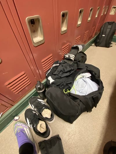 A locker room is depicted with a row of red metal lockers lining a wall, featuring recessed handles and ventilation slats. The floor is light-colored and textured, bordered by a green kick plate at the base of the lockers. In the foreground, a black duffel bag is open on the floor, containing various folded garments, including a light blue item with yellow-green drawstrings and black apparel. A pair of black Adidas slides with white stripes rests on top of the duffel bag. Adjacent to the bag, a black water bottle with a black cap stands upright. To the left of the duffel bag, a pair of black athletic shoes with white soles are on the floor, each containing a tan-colored rolled item, likely socks. Partially visible in the bottom left, a purple and green athletic shoe, possibly a cleat, is worn on a foot with a black sock. A small, circular object with a purple center is on the floor near this shoe. Further down the locker row, a black backpack leans against the wall. The scene suggests a transient state, either preparing for or concluding athletic activity in an indoor facility, possibly a school or gym in the Town of Colonie, United States. No individuals are explicitly visible, though their implied presence is indicated by the personal belongings.