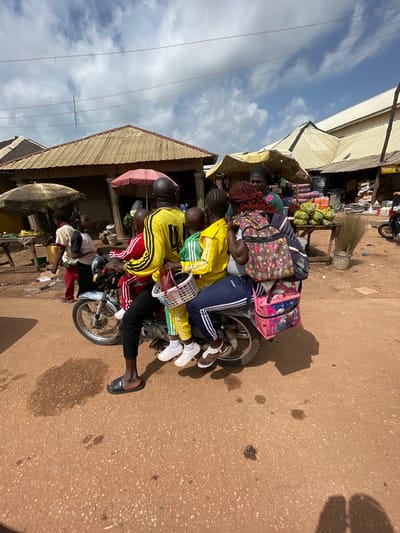 A group of people are riding on a motorcycle in a busy outdoor market in the city of Mbar, Nigeria. The driver, a man in a yellow and black shirt and black pants, is at the front. Behind him, a woman in a green shirt and a patterned headscarf is sitting. She is carrying a large patterned backpack and a pink bag with images of cartoon characters. Two children are also on the motorcycle. One child, wearing a red and white tracksuit, is sitting between the driver and the woman. Another child, in a yellow and green tracksuit, is sitting behind the woman. Both children are holding baskets. The motorcycle is parked on a dusty, reddish-brown road. In the background, there are market stalls with various goods, including produce and baskets of fruit. Several umbrellas provide shade over the stalls. The sky is partly cloudy.