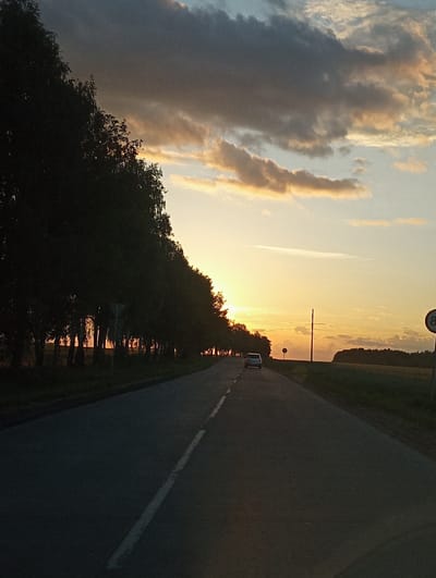 The image shows a sunset scene on a road, likely in Буйніцкі сельскі Савет, Belarus. The sun is low on the horizon, casting a warm, golden glow across the sky and illuminating the clouds with orange and yellow hues. The sky transitions from bright yellow near the sun to a lighter blue towards the left. The road is paved and has a white line marking the edge. A white car is visible on the road, moving away from the viewer.
On the left side of the road, there is a dense line of trees, silhouetted against the bright sky. Beyond the trees, a body of water reflects the colors of the sunset, creating a shimmering effect. The reflection of the trees is also visible in the water.
Further down the road, there appears to be a power line pole. A circular sign with the number "8" is visible at the top of the frame, possibly indicating a speed limit. Another smaller, circular sign is present further down the road.
The overall mood of the image is peaceful and serene, typical of a sunset. The lighting suggests it is late evening. The presence of the road, car, and utility pole indicates a developed area, likely rural or semi-rural. No people are clearly visible in the image.