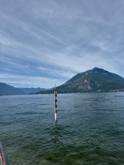 A serene lake scene unfolds under a partly cloudy sky. A marker pole, painted with alternating red and white bands, stands partially submerged in the foreground, its reflection shimmering on the water's surface. The lake stretches out towards distant, verdant mountains that frame the horizon. In the mid-ground, several small boats dot the water, suggesting leisurely activity. The overall atmosphere is one of tranquility and natural beauty, hinting at a location like Gittana, Italy, known for its picturesque lake views.