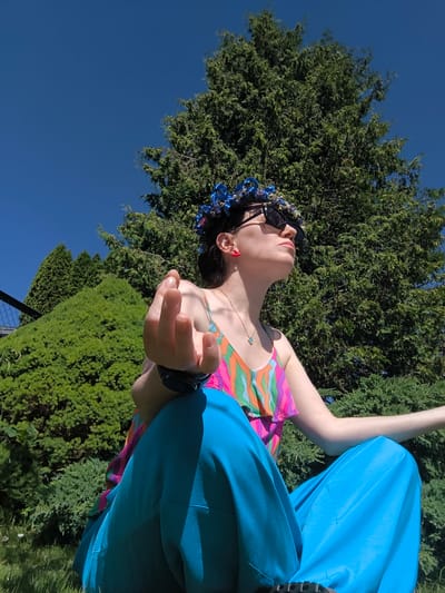 A person is meditating outdoors in Dzērumi, Latvia. They are seated on the grass, with their legs crossed in front of them, and are wearing bright, patterned clothing. They have a flower crown on their head, sunglasses, and red earrings. Their right hand is raised, with fingers extended. Behind them, a large evergreen tree and other lush greenery create a natural backdrop. The sky is a clear, vibrant blue, suggesting a sunny day. The low angle of the shot emphasizes the sky and the person's serene pose. The overall mood is peaceful and tranquil. There is no visible text in the image.