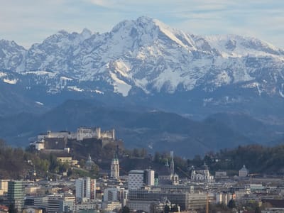 The image presents a cityscape with a mountainous backdrop. In the foreground, there is a dense urban area with buildings of varying heights and architectural styles. A historical fortress or castle sits atop a hill in the middle ground, surrounded by trees. Behind the city and castle, a series of mountains are visible, with the highest peaks covered in snow. The sky is partly cloudy.