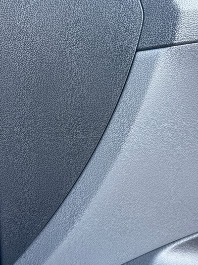 The image shows a close-up view of a textured, matte plastic surface with two distinct color sections and a curved seam. The left section is a darker gray, and the right section is a lighter, silvery gray. The texture on both sections appears to be a uniform, fine-grained pattern, possibly resembling dimpled leather or a similar synthetic material. The lighting suggests a bright environment, casting subtle shadows and highlighting the texture. There are no people, animals, or other objects visible in the frame. The context of Parekklisia Community, Cyprus, is not visually evident from the image itself, as it appears to be a detail shot of a material rather than a scene from a specific location.