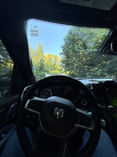 This media file is a first-person perspective shot from inside a vehicle, likely a Ram truck, as indicated by the prominent Ram logo on the steering wheel. The driver's seat is visible, with the steering wheel, dashboard, and part of the center console in the foreground. The steering wheel has various controls, including buttons for cruise control and audio. A phone mount is also visible on the dashboard. Through the windshield, a bright, clear sky is visible, suggesting daytime. Lush green trees and foliage surround the vehicle, indicating an outdoor setting, possibly a residential area or a park. One of the windshield stickers, a Jiffy Lube service reminder, is partially visible with text that includes "Service Oil Change," "67502 MILES," and "10/3/2025." The image appears to be taken on a sunny day. There are no people visible in the image.