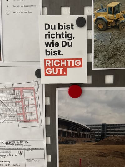 The image depicts a close-up view of a wall or bulletin board with various documents and a large photograph attached. In the upper left, a snippet of a document with German text is visible, mentioning "Kontroll- und Spül-schacht neu" (Control and flushing shaft new) and "neu zu pflanzender Baum" (new tree to be planted).

Dominating the center of the image is a white card with bold German text that reads "Du bist richtig, wie Du bist. Richtig. GUT." (You are right as you are. Right. GOOD.). This card is secured by a black circular magnet.

To the right of the text card, a portion of a photograph shows a yellow Volvo wheel loader on a muddy construction site.

Below the text card and to the left, a blueprint or architectural drawing is visible, showing a grid-like layout with street names like "Planstraße B" and "Planstraße C". The drawing is labeled "BLATT 2002". A red circular magnet is attached to the bottom edge of this blueprint.

Further down, the lower left section of the image displays the letterhead of "SCHERER & KURZ Beratende Ingenieure" (Consulting Engineers), indicating services such as "Verkehrsanlagen • Wasserwirtschaft • Vermessung • Geoinformation" (Traffic facilities, Water management, Surveying, Geoinformation). Contact details include a phone number and email address, with a location potentially linked to Hohenbrunn, Germany, given the text "Hohenbrunn". The document also mentions "Ismaning" and "Ergänzt am:" (Supplemented on:) with a date "21.09.2016".

To the right of the blueprint and letterhead, a large photograph shows a construction site with a partially built structure featuring a pitched roof and multiple stories of windows. Piles of dirt and debris are scattered across the foreground. The sky above is overcast.