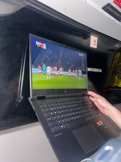An open black HP Omen laptop displays a football match, with players lined up on a green field. The laptop sits on a reflective black surface with "EMERGENCIA" printed in white. A hand partially visible near the top right edge of the laptop. In the upper left corner, a grey rectangular object with small, dark square details is visible. The background includes blurry grey and gold elements.
