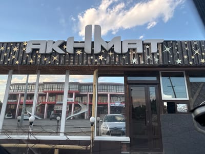 This image shows the exterior of a building with a prominent sign that reads "AKHMAT". The sign is made of large, white, three-dimensional letters and is mounted on a dark wooden facade decorated with numerous golden starbursts. The building itself appears to be a multi-story structure with a red and white color scheme. Reflective glass windows on the facade show reflections of the building's interior, including staircases and vehicles.

In the foreground, a silver car is visible through the glass. The sky above is blue with scattered white clouds, suggesting it is daytime. The overall impression is of a commercial establishment, possibly a restaurant or shop, given the signage and the building's design. The location context indicates this is in Kurchaloy, Russia. No people are clearly visible in the image, and the scene appears calm and still. The visible text includes the building's name and some Russian signage within the reflections, such as "ЭФАН АППО-ТАМХА" which translates to "Cafe Appo-Tamkha."