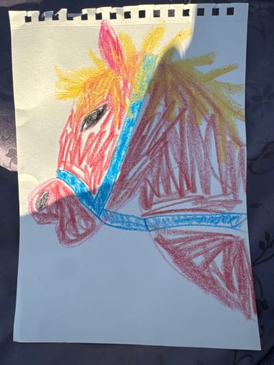 The image presents a hand-drawn illustration, likely by a child, depicting the head and upper body of a horse in profile, oriented towards the left. The drawing is executed on a rectangular sheet of white paper, approximately A4 size, which exhibits a perforated top edge, indicating detachment from a spiral-bound notebook. The horse is predominantly colored with red crayon or pastel, featuring a spiky, yellow-orange mane and a distinct blue bridle wrapping around its muzzle and extending across the upper neck area. A black eye and nostril are visible on the horse's head. The paper rests upon a dark, subtly patterned surface, possibly a fabric tablecloth. Strong, directional natural light illuminates the scene, casting prominent shadows: a diagonal shadow partially obscures the horse's mane and upper back on the right side of the drawing, and another shadow borders the paper's left edge. The location of this scene is Saint-Mandé, France.