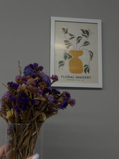 The image shows a framed print on a gray wall. The print features a graphic design of stylized green leaves and a mustard-yellow vase-like shape. Text reading "FLORAL IMAGERY ARTWORK SINCE 1921" is printed on the right side of the artwork. In the foreground, there is a vase of dried flowers with purple and blue blooms, and a hand with a blue-tipped nail touches the vase. The location is not discernible from the image, and therefore it's impossible to confirm if it's Zagarancea, Moldova.