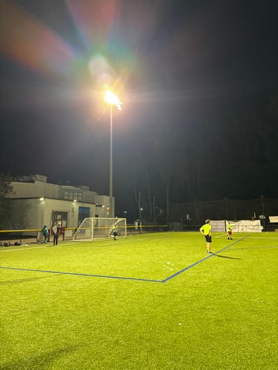 The image depicts an outdoor artificial turf sports field at night in Newton, United States. The field is green with visible white and blue markings. A white soccer goal with a net is positioned on the left side of the field. A tall stadium light pole stands prominently on the left, emitting bright yellow-white light that creates significant lens flare effects across the upper left quadrant of the dark night sky.
In the background on the left, a light-colored, two-story building with multiple windows is visible. Dark trees and what appears to be a tall perimeter fence or netting line the far background to the right and behind the building.
Four individuals are present on the field. One male in a bright yellow short-sleeved shirt and dark shorts stands centrally in the mid-ground, facing right with his hands on his hips. Another male, positioned near the white goal as a goalkeeper, wears a yellow-green long-sleeved shirt and dark pants, facing right. Two additional individuals are partially visible further to the left, near the building, one wearing a light blue/green hooded jacket; they are standing stationary. A fourth individual, wearing a red shirt, is partially visible further downfield to the right of the central yellow-shirted person. The scene suggests a nighttime sports activity, likely soccer.