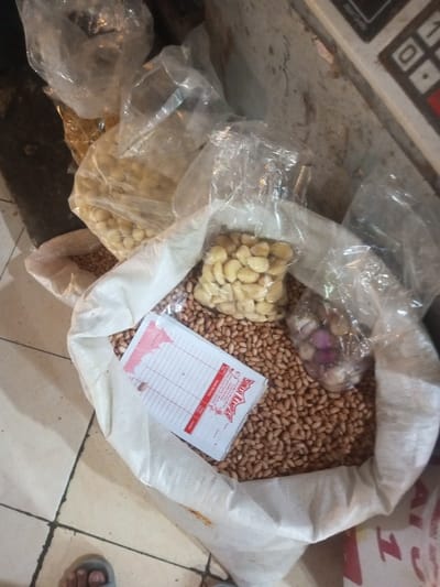 A top-down shot reveals a white sack filled with peanuts. Scattered atop the peanuts are plastic bags containing garlic cloves and shallots. A receipt with red trim sits on the peanuts within the sack. Additional bags of garlic and what appears to be nuts are visible around the sack. A corner of a weighing scale is visible in the upper right, with tiled flooring in the lower left.