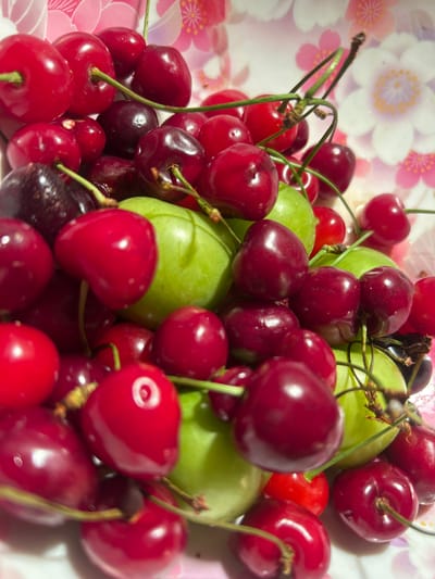 The image shows a close-up of a bowl filled with red cherries and some green grapes. The cherries vary in shade from bright red to deep burgundy, and some have stems attached. The green grapes are round and have a smooth skin. The background is a blurry pink and white floral pattern, likely the design of the bowl or tablecloth. The lighting suggests an indoor setting. There are no people or animals visible in the image. The scene is a still life of fruit. The location in Getapnya, Armenia is not determinable from the image alone.