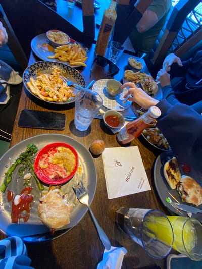 The image captures a table setting, presumably in a restaurant or pub, featuring several diners enjoying a meal. The location is possibly in Elmbridge, United Kingdom.
The table is wooden, with various plates of food, glasses of beverages, and other dining accessories scattered across it. The meals consist of fries, burgers, and what appears to be a pasta or noodle dish. Plates contain both untouched food and remnants of what has been consumed. Condiments, including a bottle of sauce and small bowls of ketchup, are also present.
Diners' hands and partial bodies are visible around the table, suggesting an ongoing dining experience. Some diners appear to be interacting with their food, while others are engaged in conversation or other activities not fully captured in the image. A napkin and a mobile phone are also on the table.