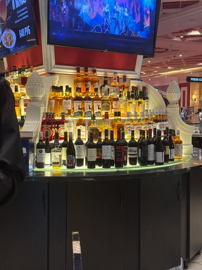 The image captures a bar setting, likely in Shkotovo, Russia, as indicated by the Russian text on a nearby screen displaying "640 РУБ", which translates to "640 Rubles", next to food images. The bar features a lavish display of various liquor and wine bottles arranged on illuminated shelves behind the counter. Brands like Chivas, Black Ram, The Whistler, and Jim Beam are visible, along with Russian labels such as “КРУТОЯР” (Krutoyar). A large TV screen is mounted nearby, showing a vivid, theatrical performance, potentially related to a show called "WEB". The bar is partially obscured by a dark-clad person, presumably a patron, and the countertop, made of what appears to be a sleek black material. The background shows ornate decorative elements and a sign saying "POKER CLUB".