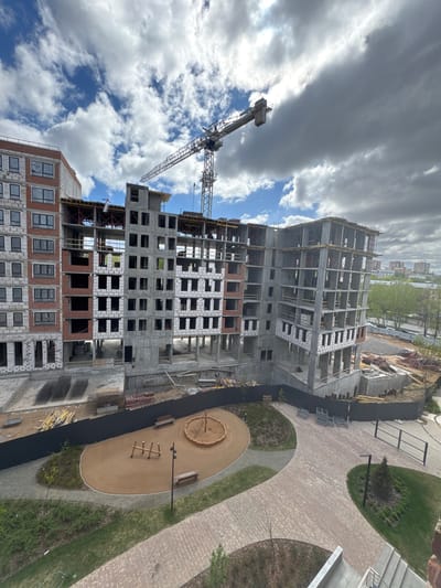 The image depicts a construction site in Люк, Russia. A high-rise building is under construction, with a crane nearby. The building is in its skeletal form, with concrete and brick work visible. A playground with sand, benches, and climbing structures is adjacent to the construction site. The sky is cloudy with patches of blue.